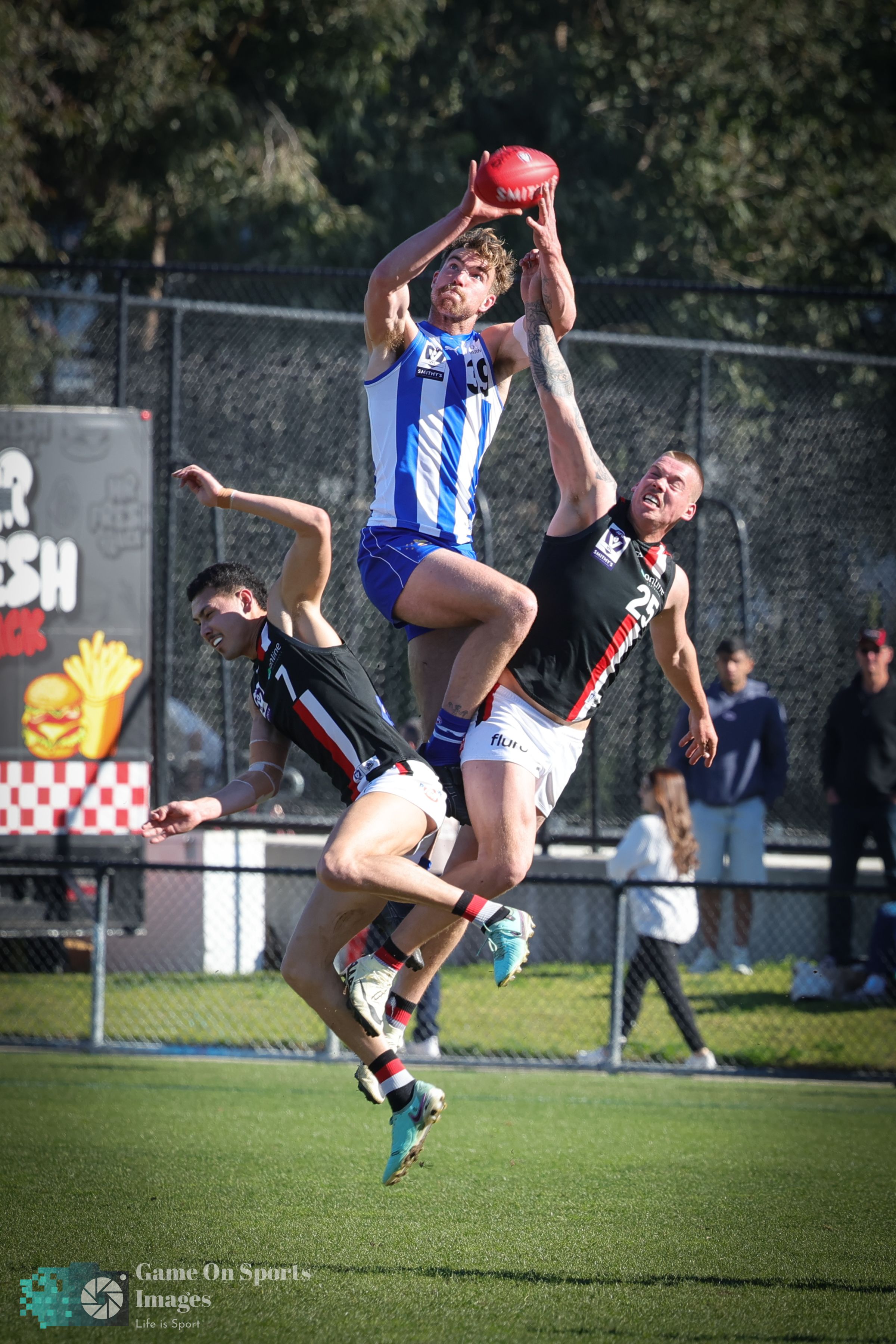 Athletes jump and compete for possession of the ball during an intense Australian Rules Football match on a sunny day.