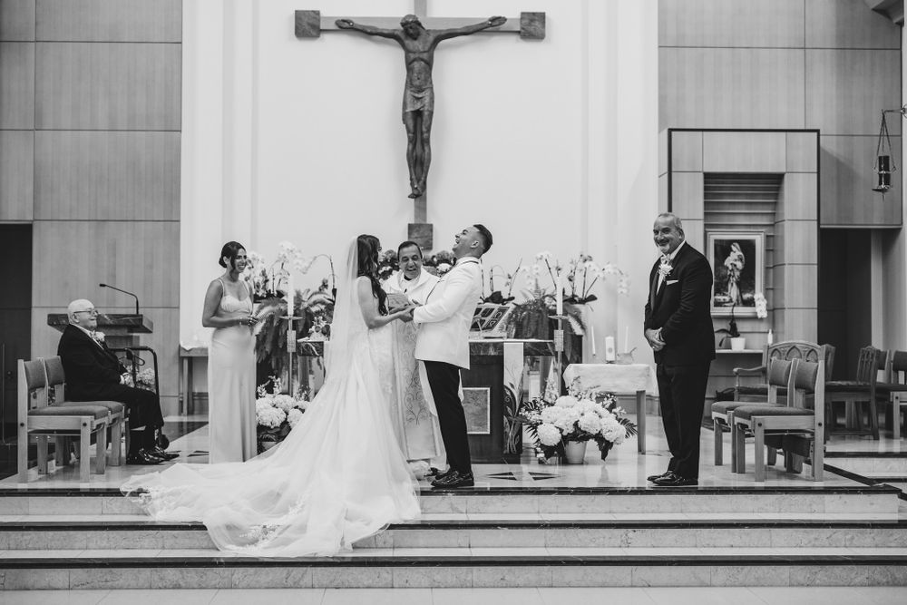 A black and white wedding ceremony photo in a church with a large crucifix mounted on the wall.
