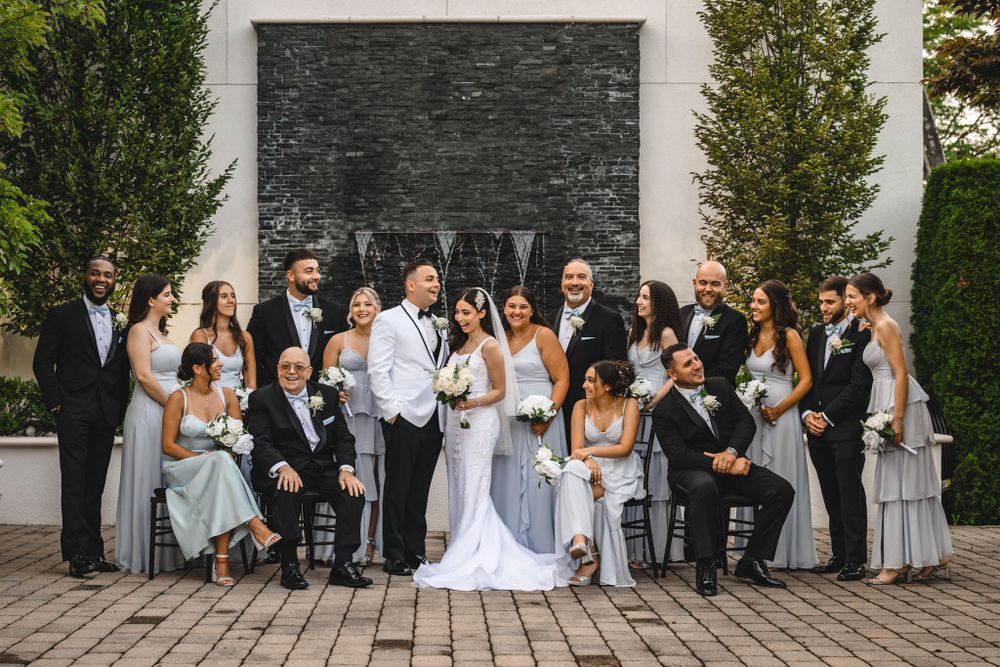 Wedding party group photo against stone wall backdrop with trees and landscaping.