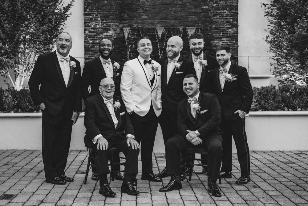 Groomsmen in formal black suits pose together outdoors against a brick wall.