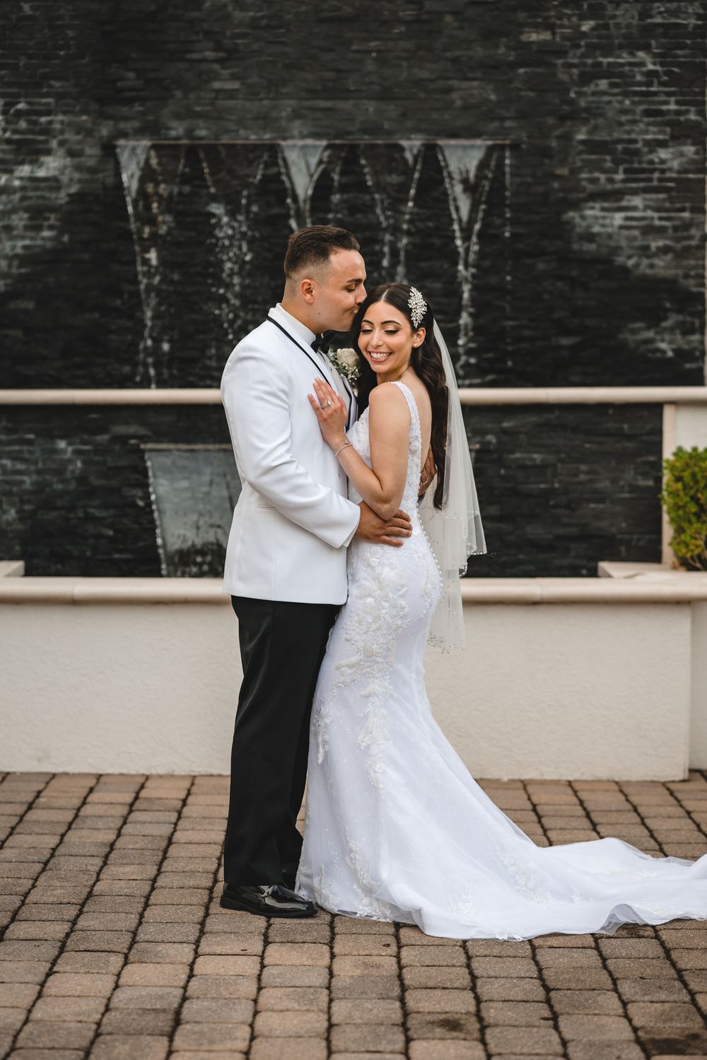 Wedding couple embraces on brick patio with bride wearing lace gown and long veil.