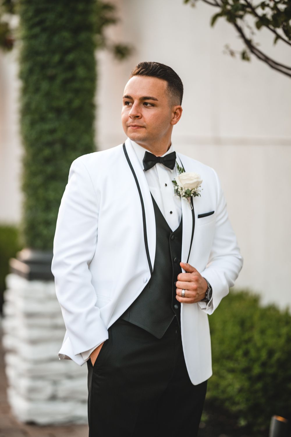 Groom in white tuxedo jacket and black bow tie poses against garden backdrop.