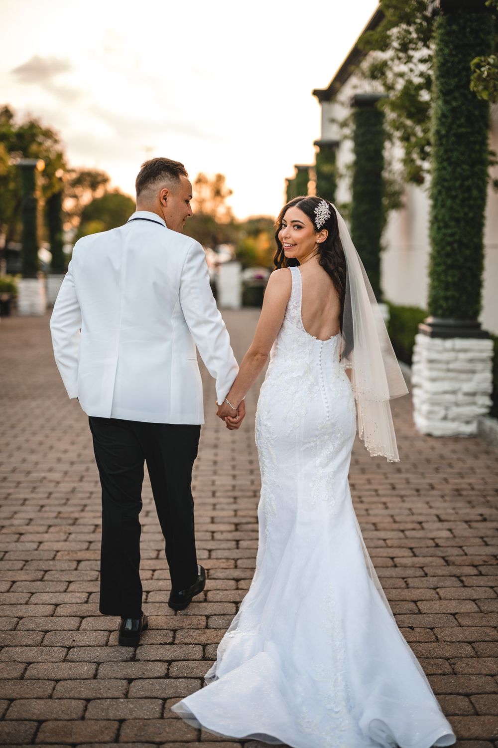Bride in lace gown and groom in white jacket walk together on brick pathway.