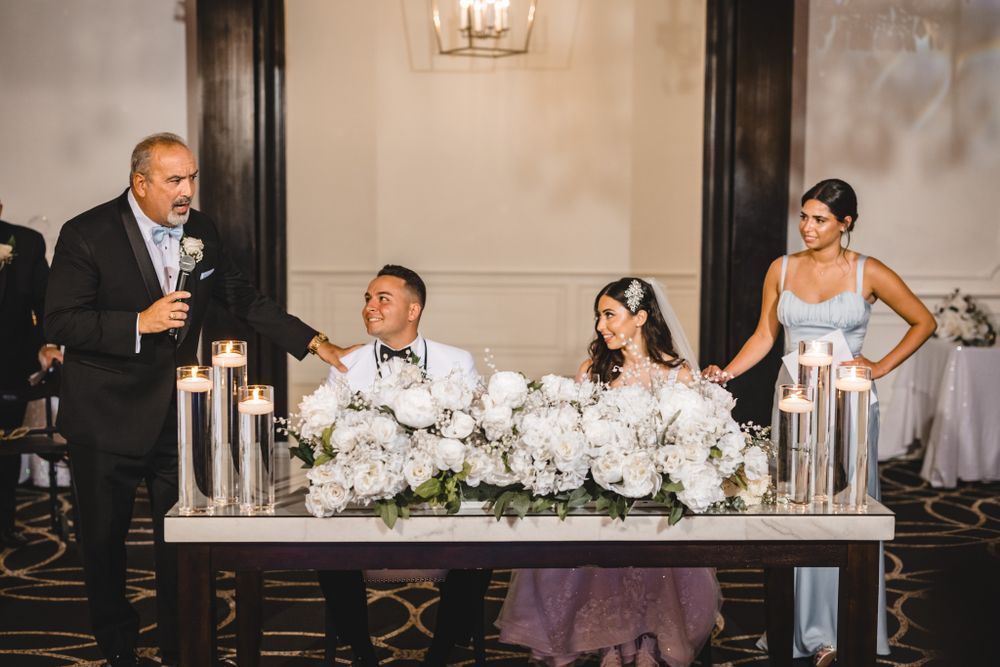 Wedding reception speech at head table decorated with white floral arrangements and elegant candles.