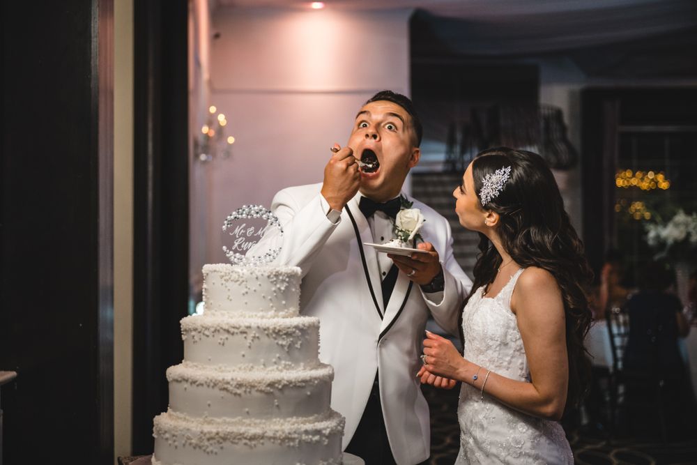 Wedding celebration with a white tiered cake visible in the background as guests dance and celebrate.