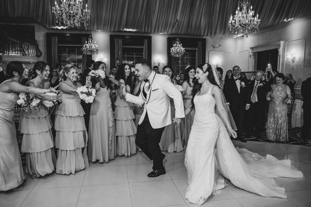 Wedding party dancing in a grand ballroom with bridesmaids in matching grey dresses and elegant chandeliers.