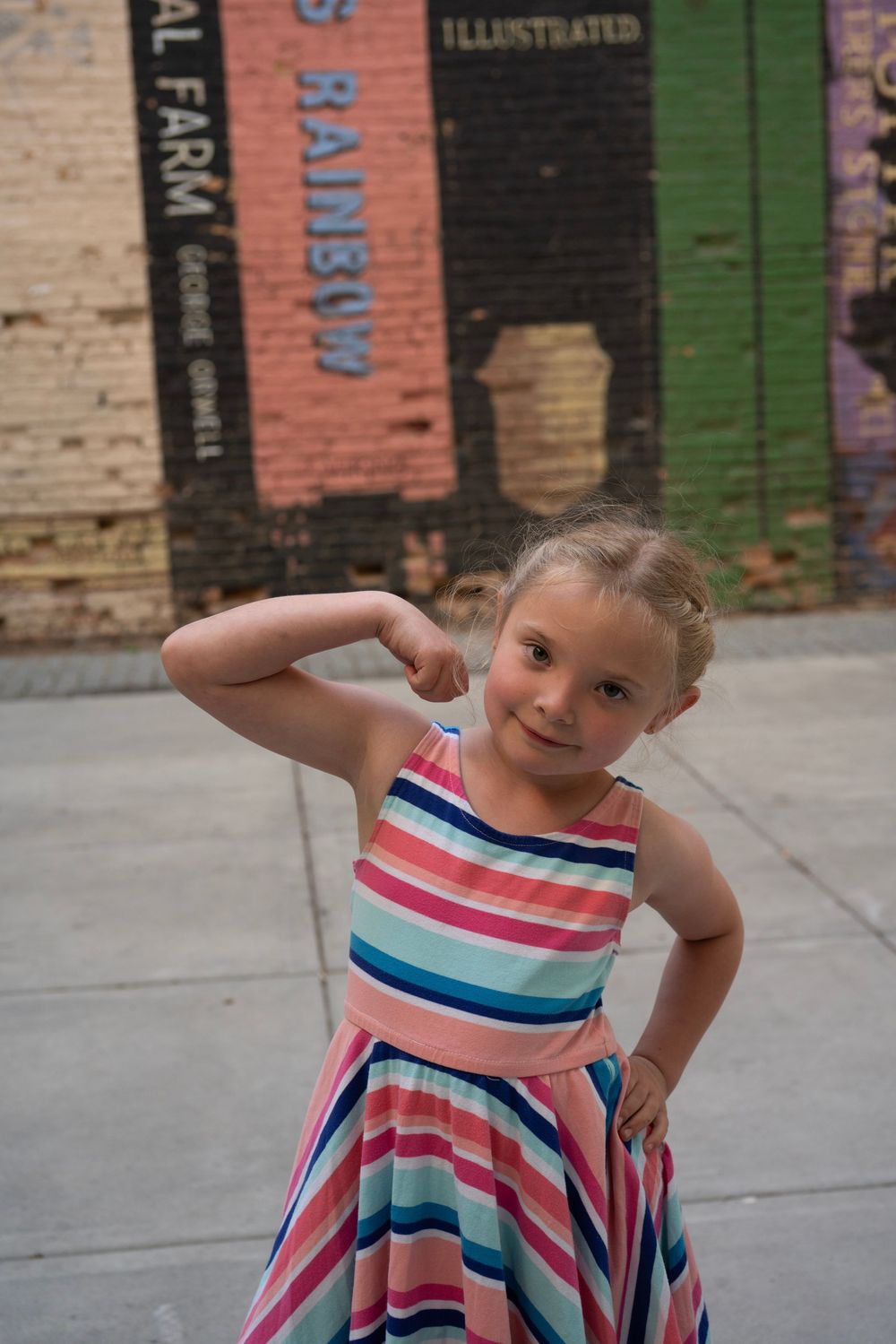 A young girl in a colorful striped dress flexes her muscles in front of a vibrant mural on a brick wall.