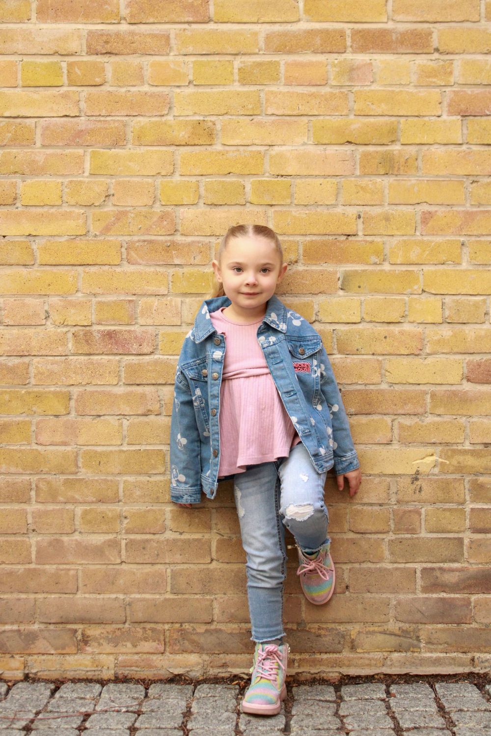 Child in denim jacket and pink top posing casually against yellow brick wall.