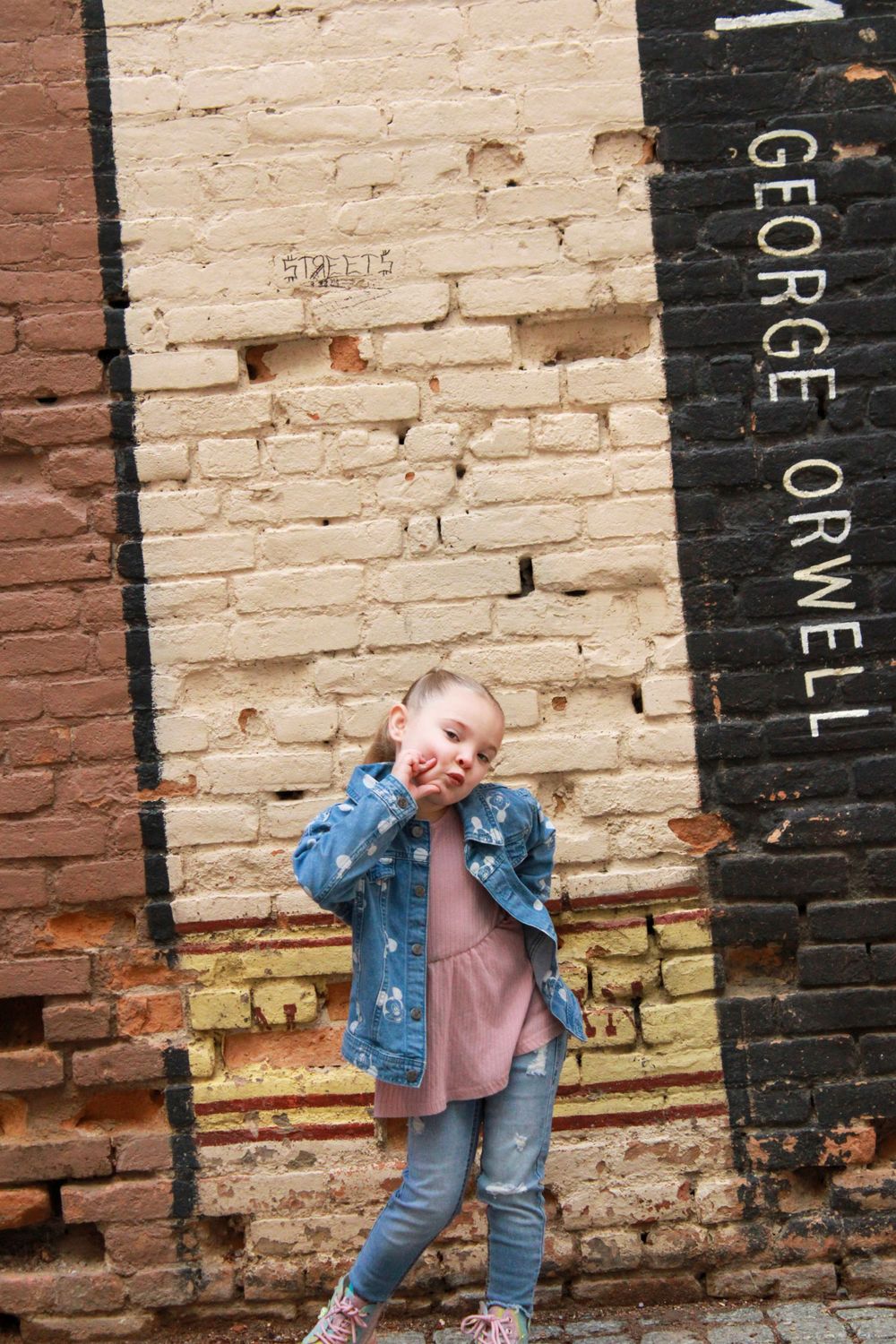 Child in denim jacket posing playfully against a brick wall.