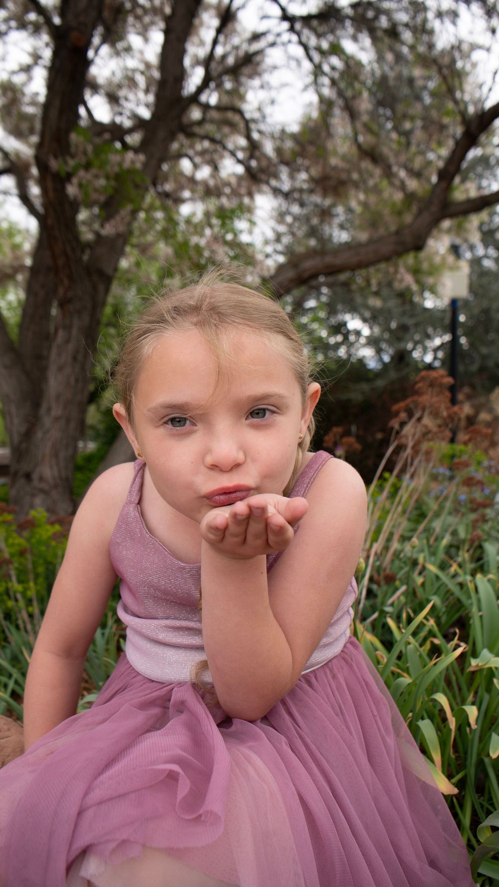 Young girl in pink tulle dress blowing a kiss outdoors in a garden setting with trees in background.