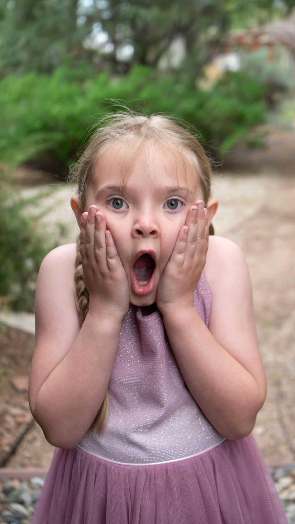 A surprised young girl in a pink dress holds her face with both hands, mouth wide open, standing outdoors.
