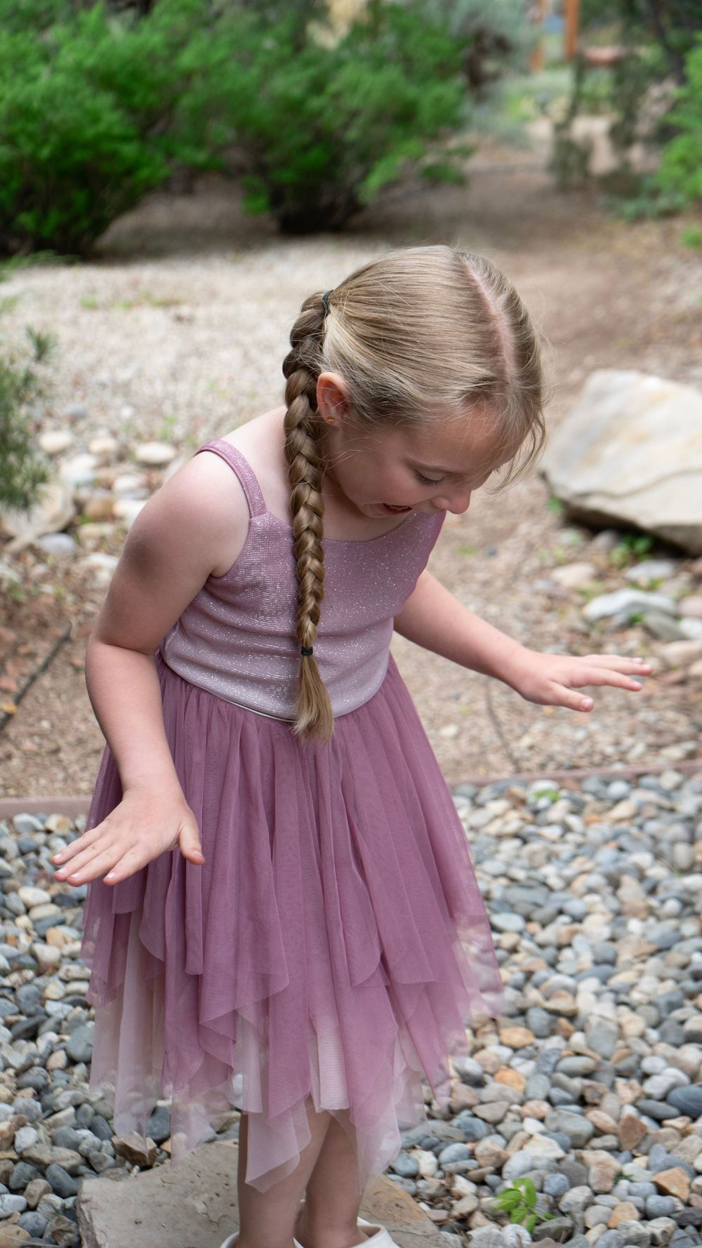 Young girl in pink tulle dress with braided hair plays on a rocky garden path outdoors.