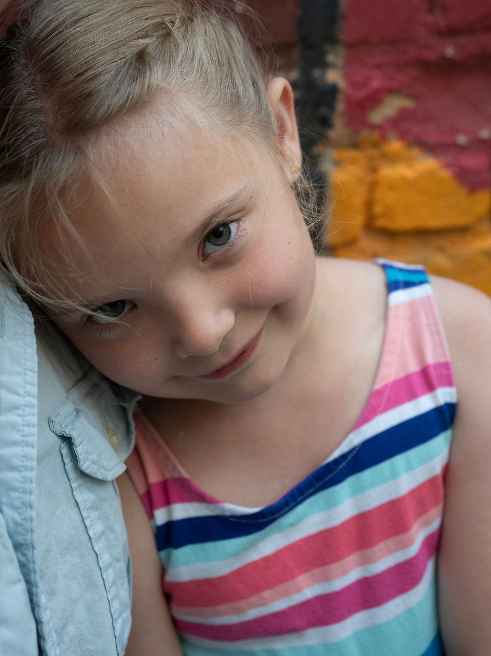 Young girl in striped tank top leaning against wall, smiling softly with colorful brick background.