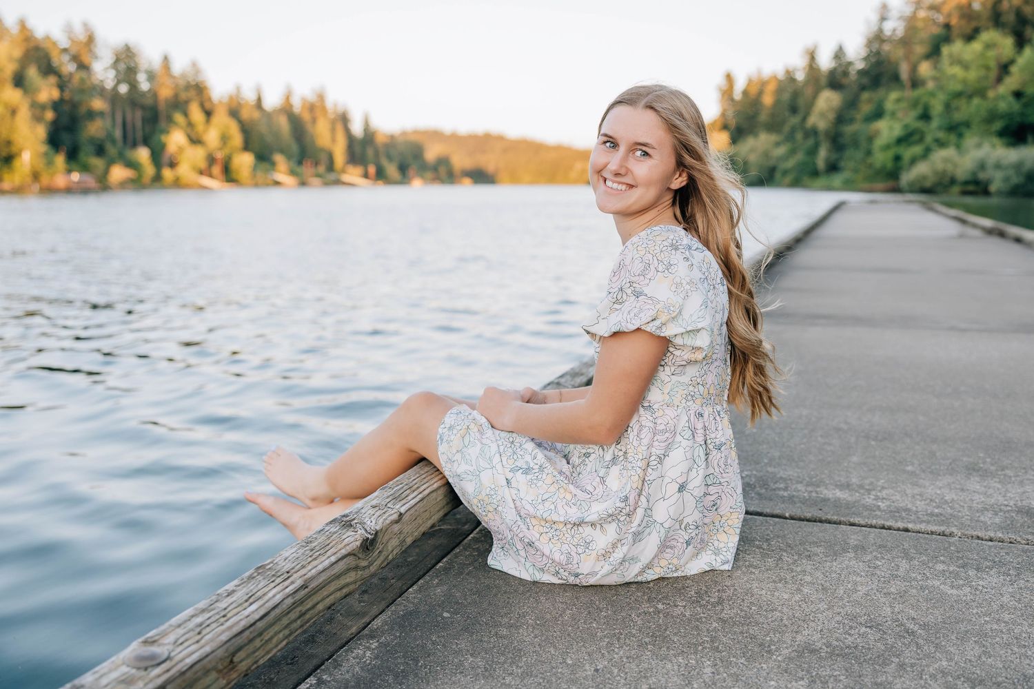 Smiling blonde girl in floral dress sits barefoot on wooden dock by serene lake.