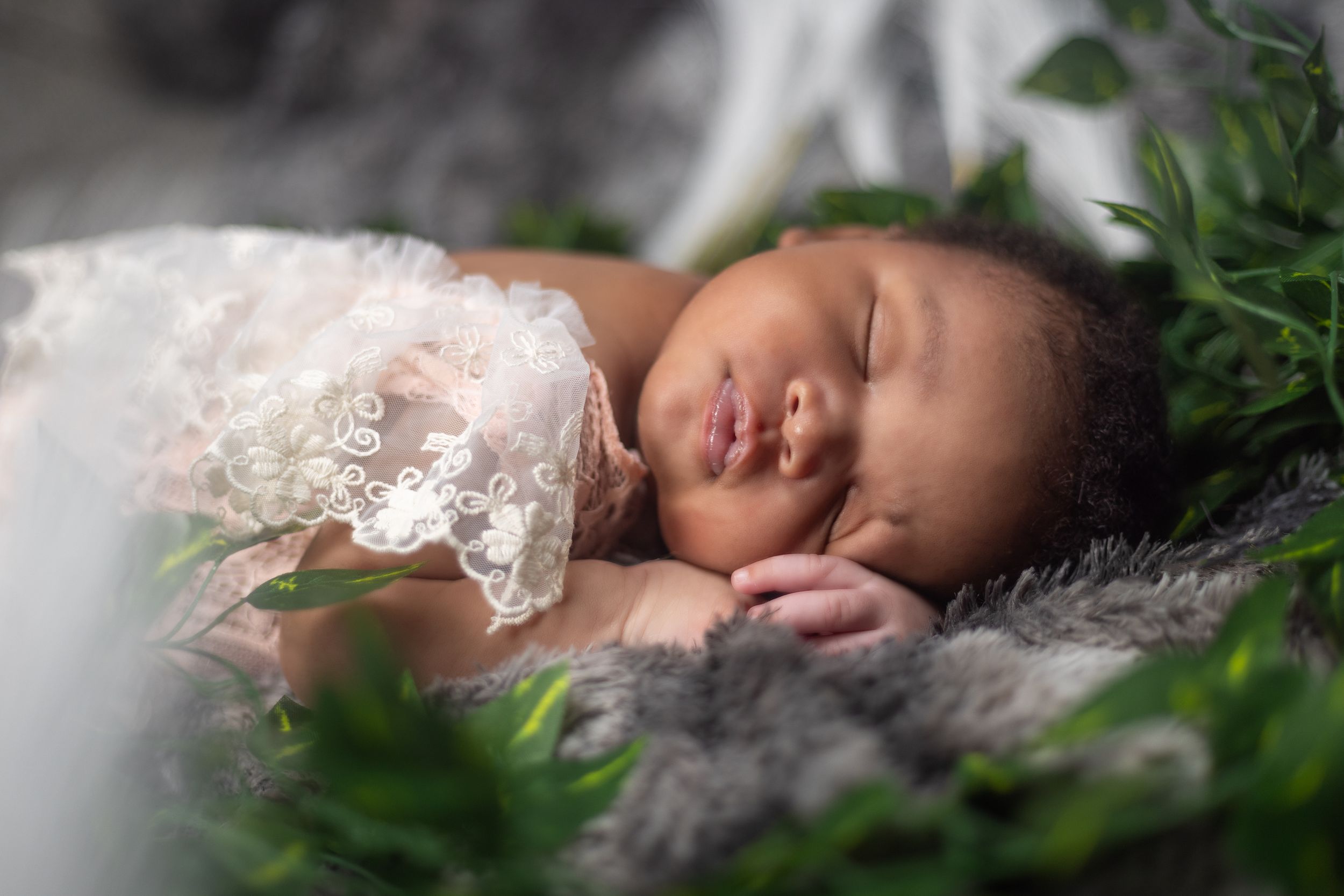 Peaceful newborn sleeps on soft blanket surrounded by greenery and lace.