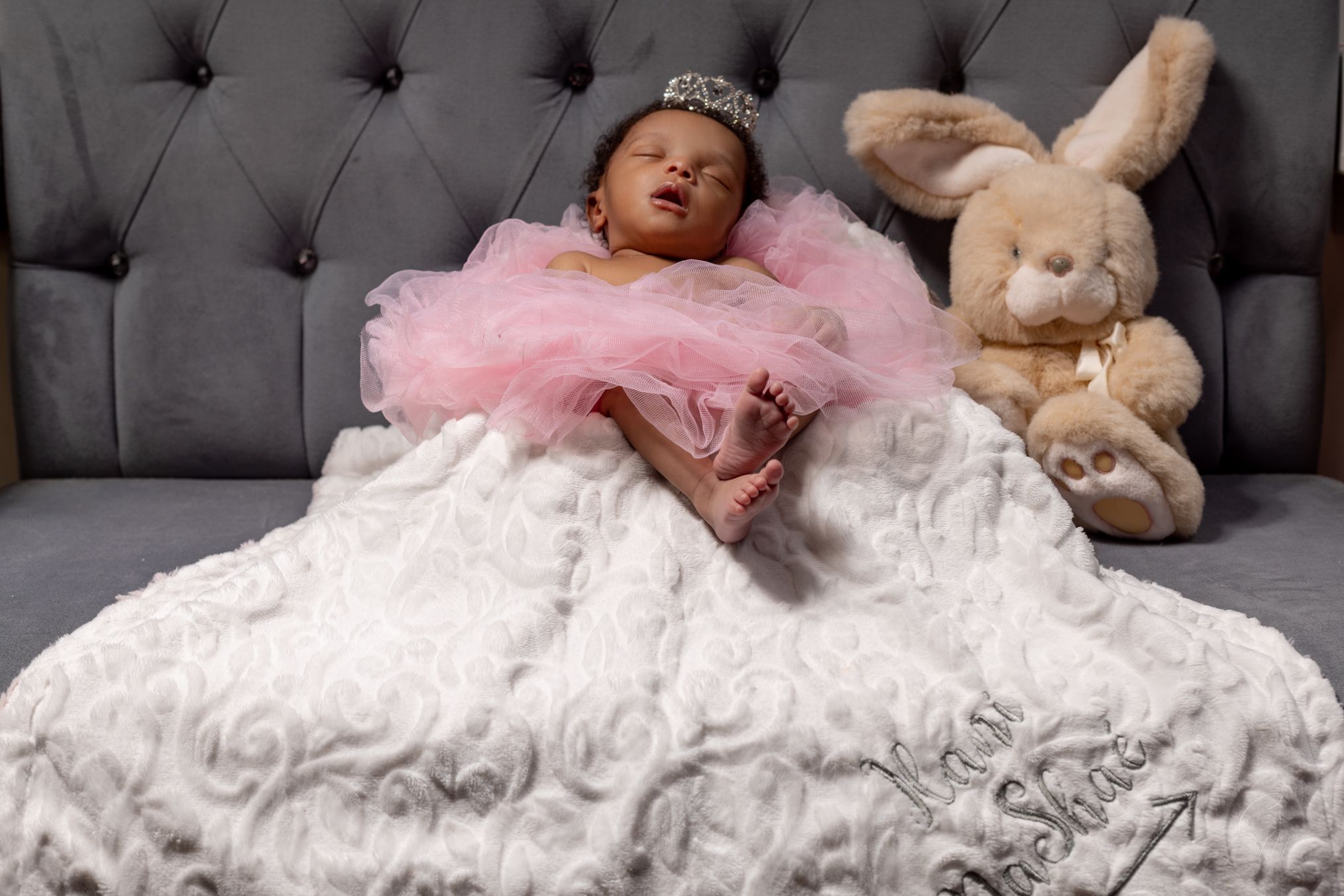 Baby sleeping peacefully in a pink tutu dress on a gray couch next to a plush bunny toy.