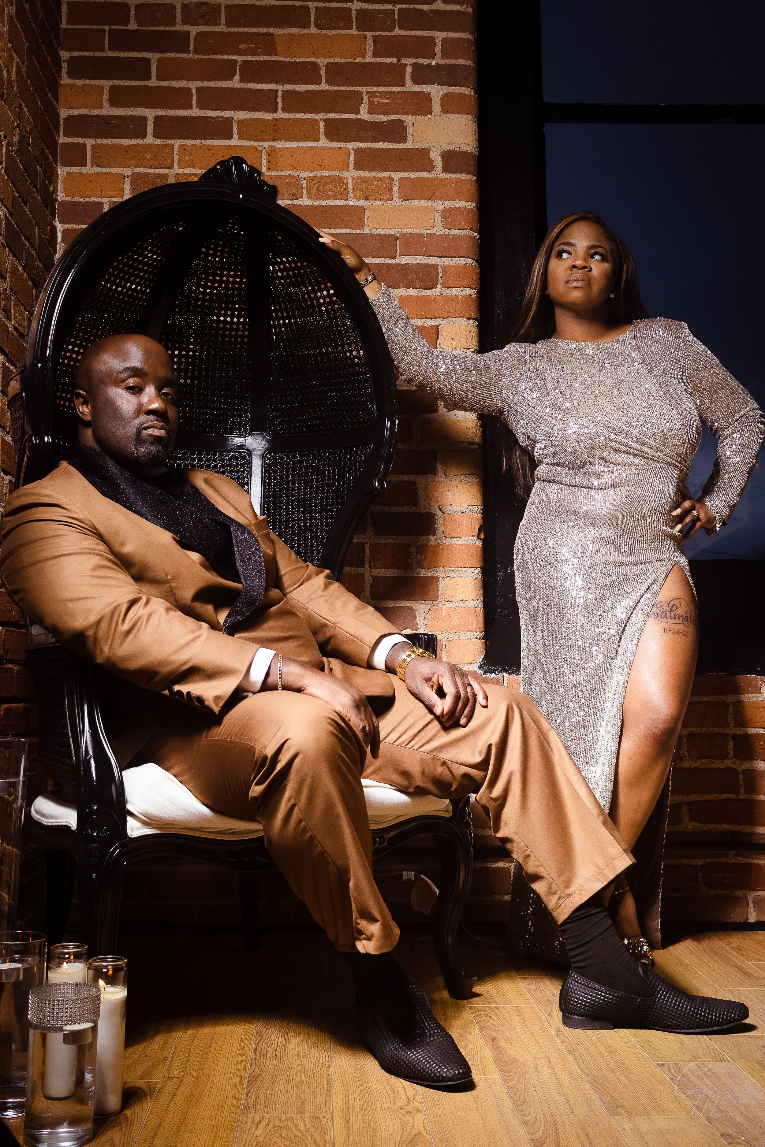 Stylish couple poses against brick wall, wearing formal bronze suit and sparkly silver dress.