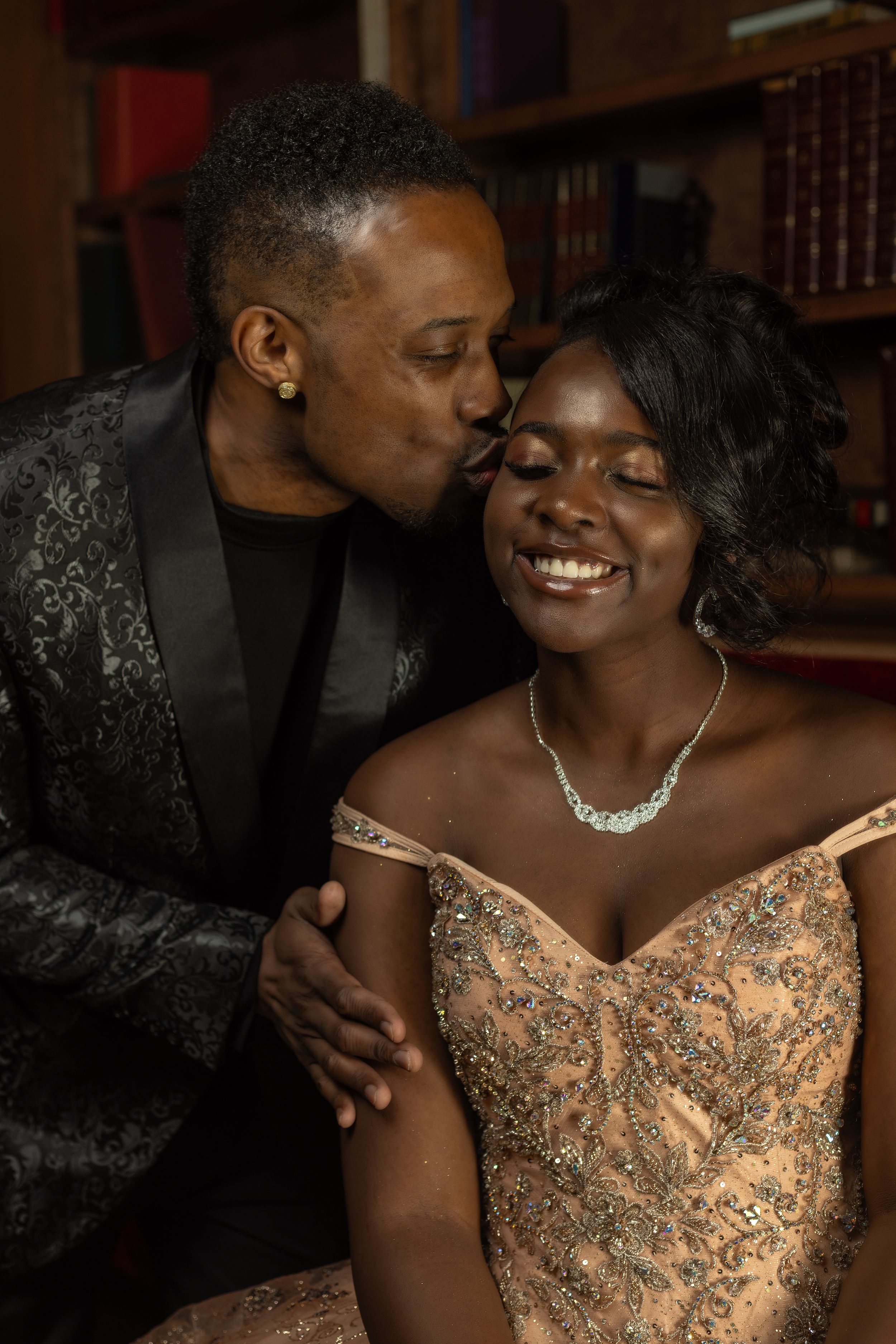 A romantic couple shares an intimate moment, with one person wearing a sparkly dress and necklace while the other wears a dark blazer.