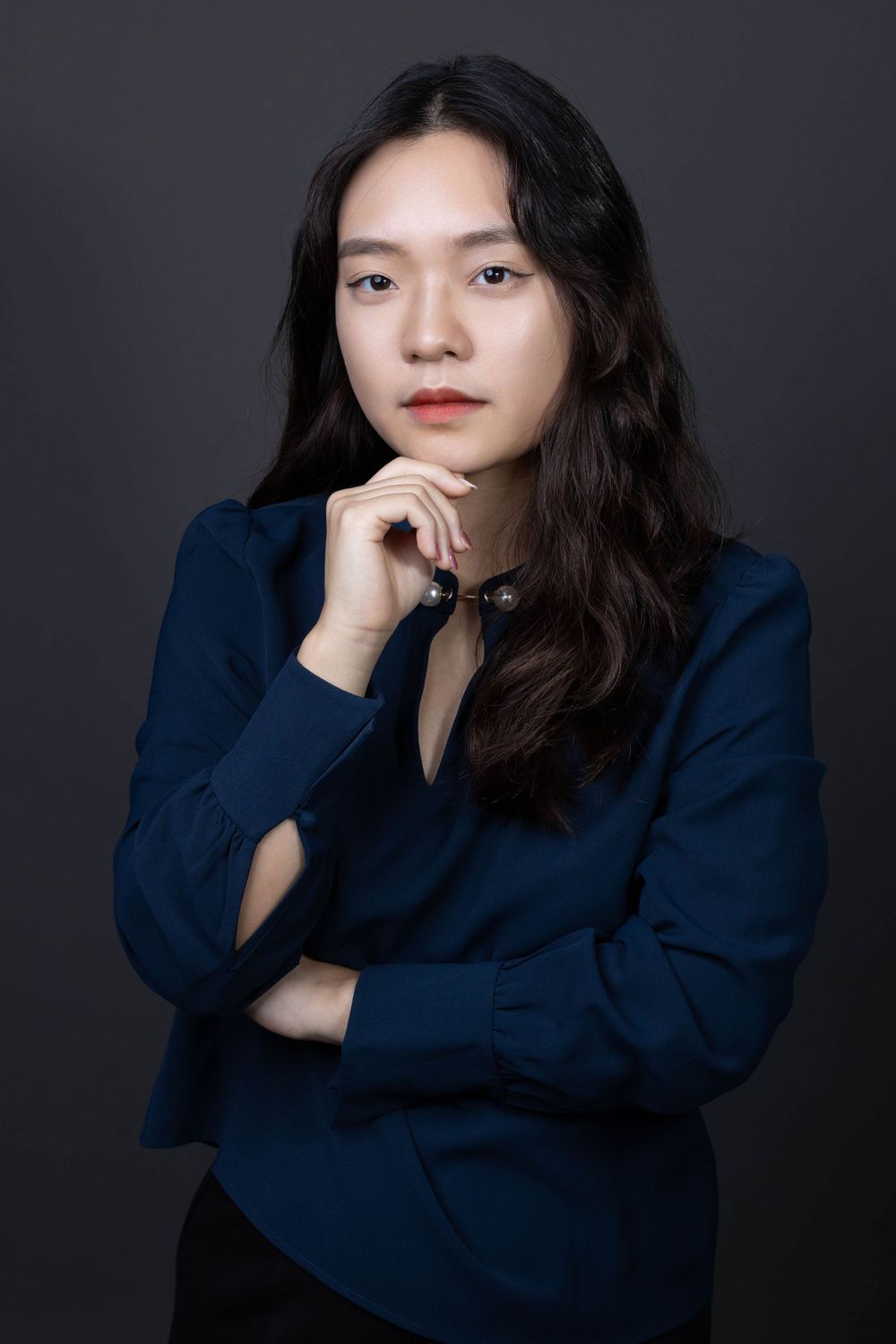 Young Asian woman in navy blazer posing thoughtfully with hand on chin against dark background.