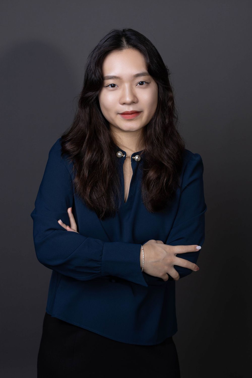 Professional Asian woman in navy blouse standing with arms crossed against dark studio background.