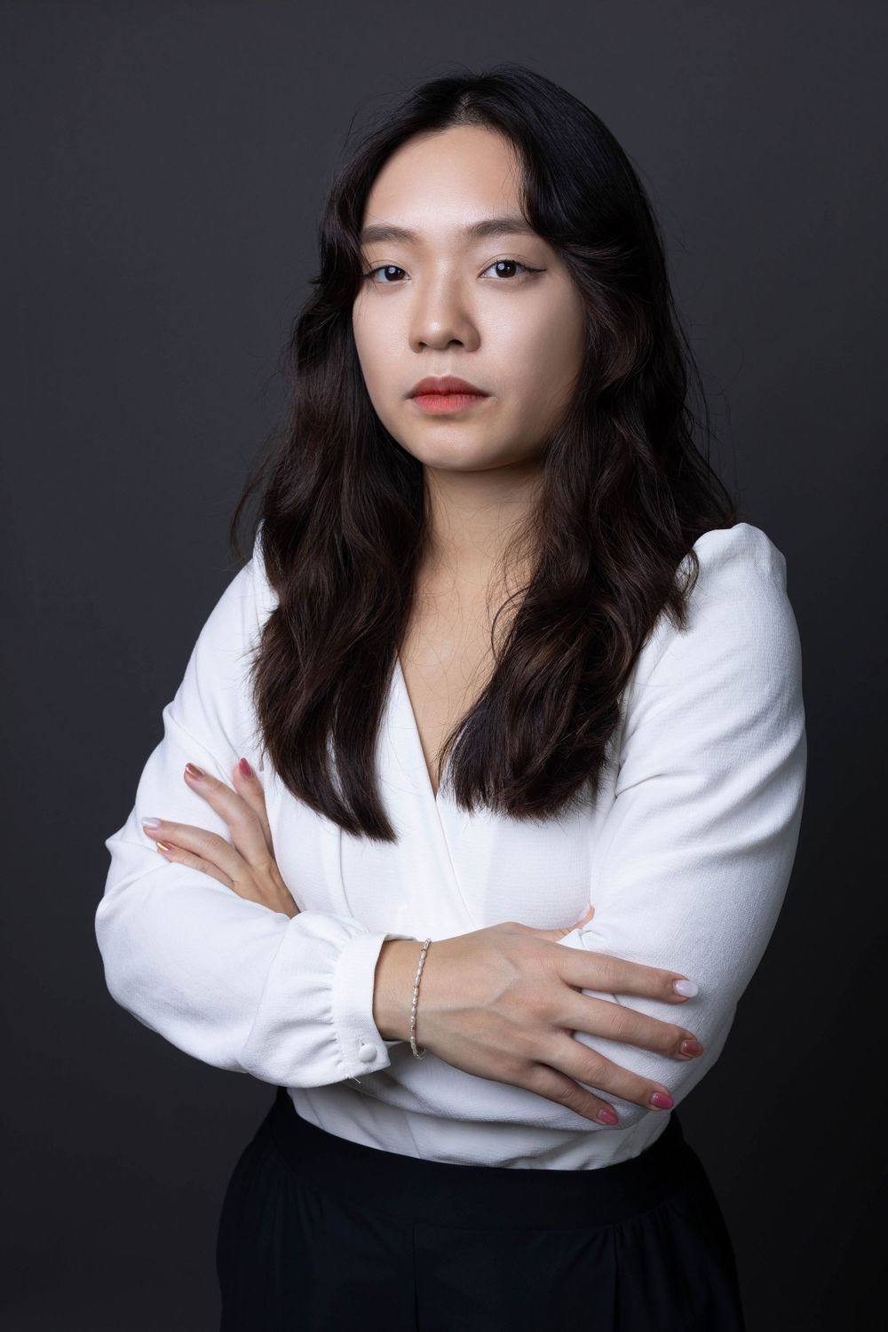 Confident Asian woman in white blouse with arms crossed posing against dark gray background.