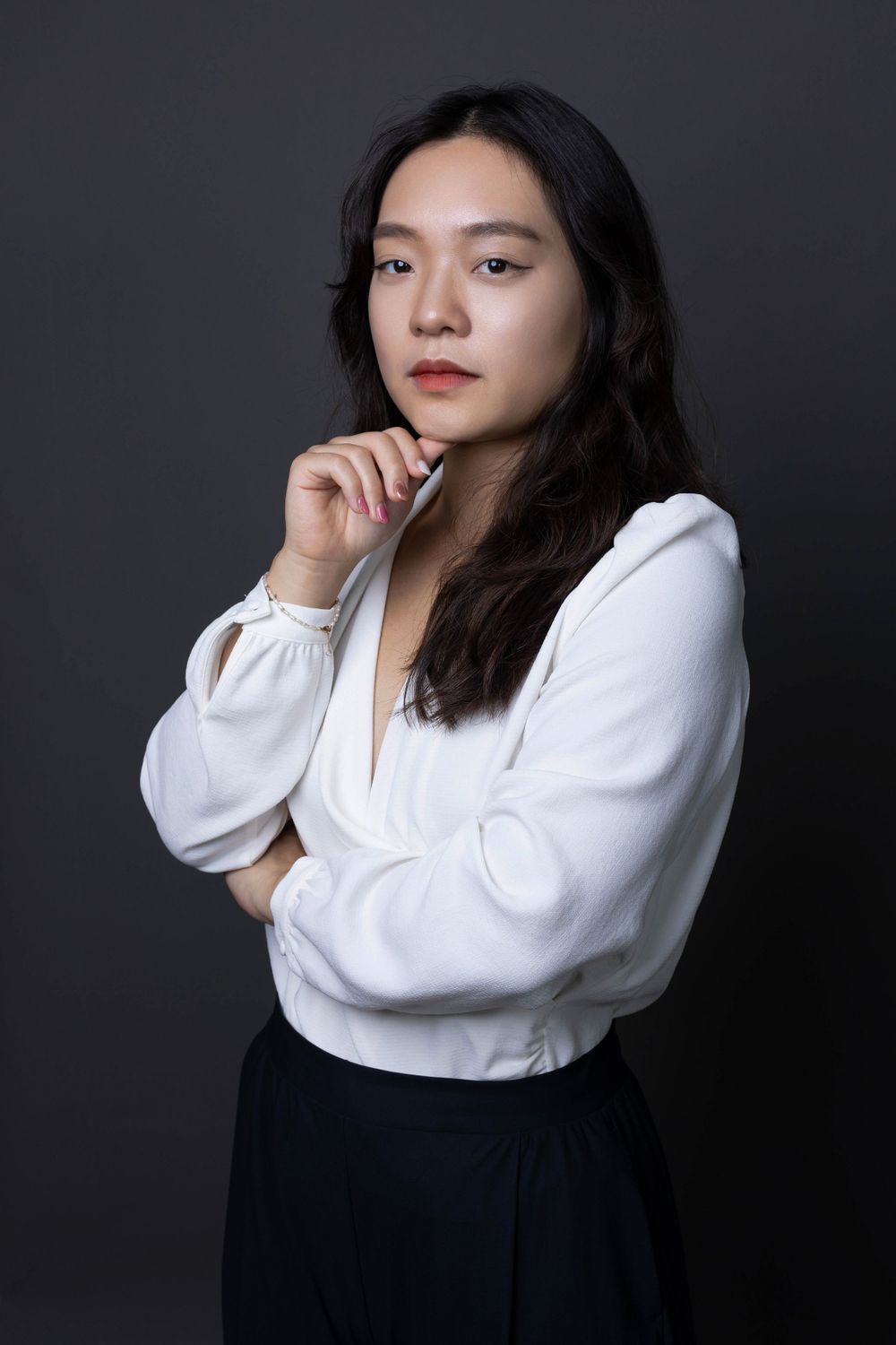 Thoughtful Asian woman in white blouse posing with hand on chin against dark studio background.