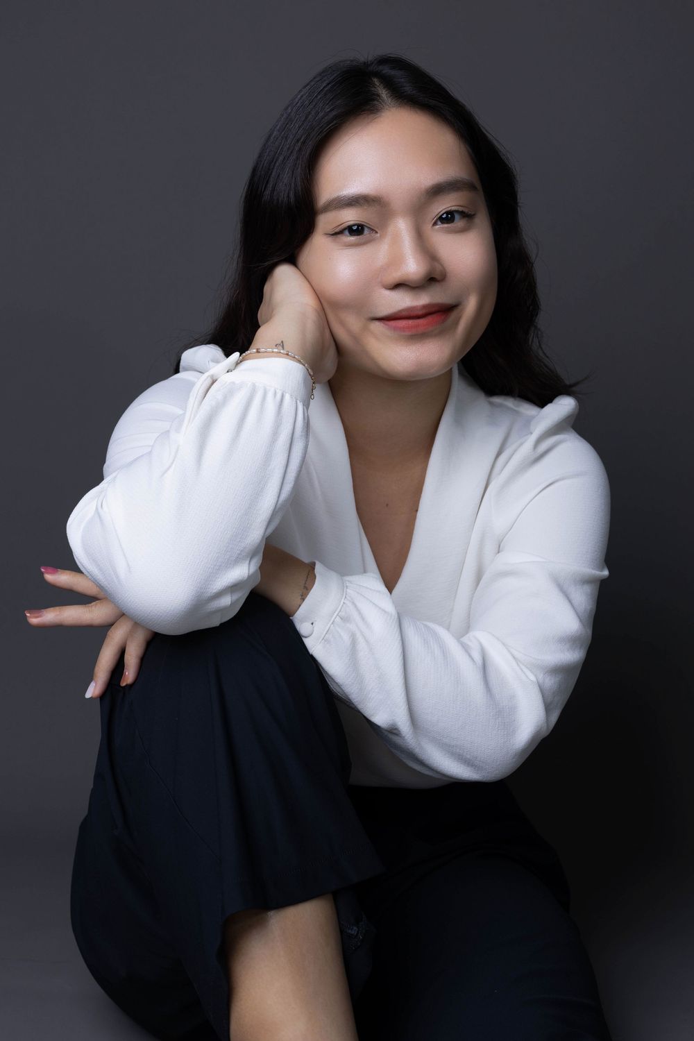 Smiling Asian woman in white blouse sitting relaxed with hand on cheek in studio portrait.