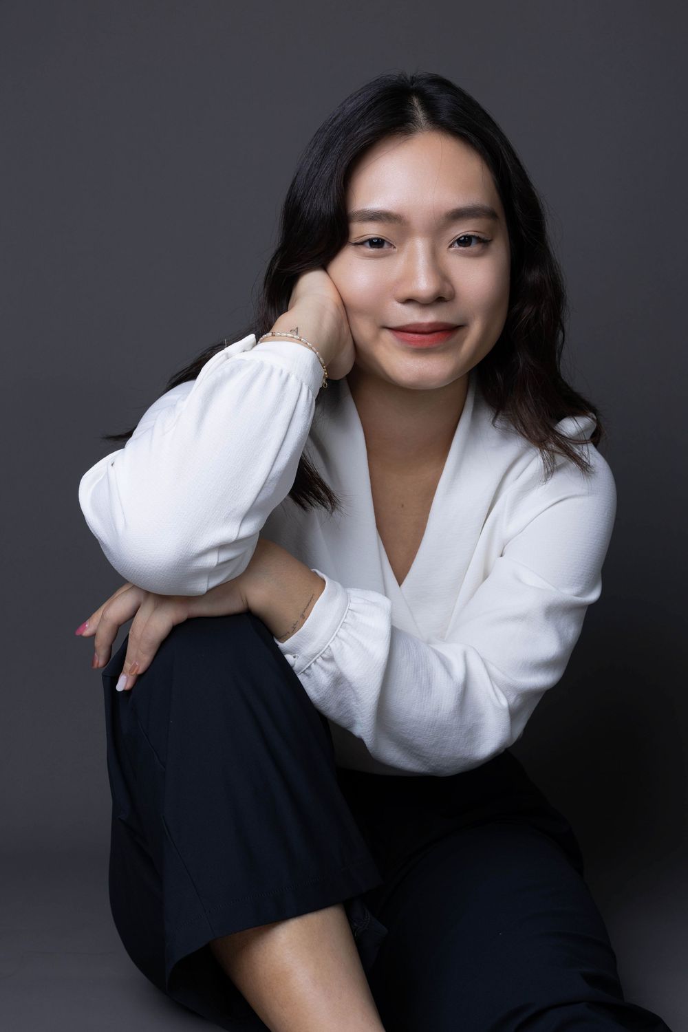 Asian woman in white blouse sitting casually with hand on cheek smiling in studio portrait.