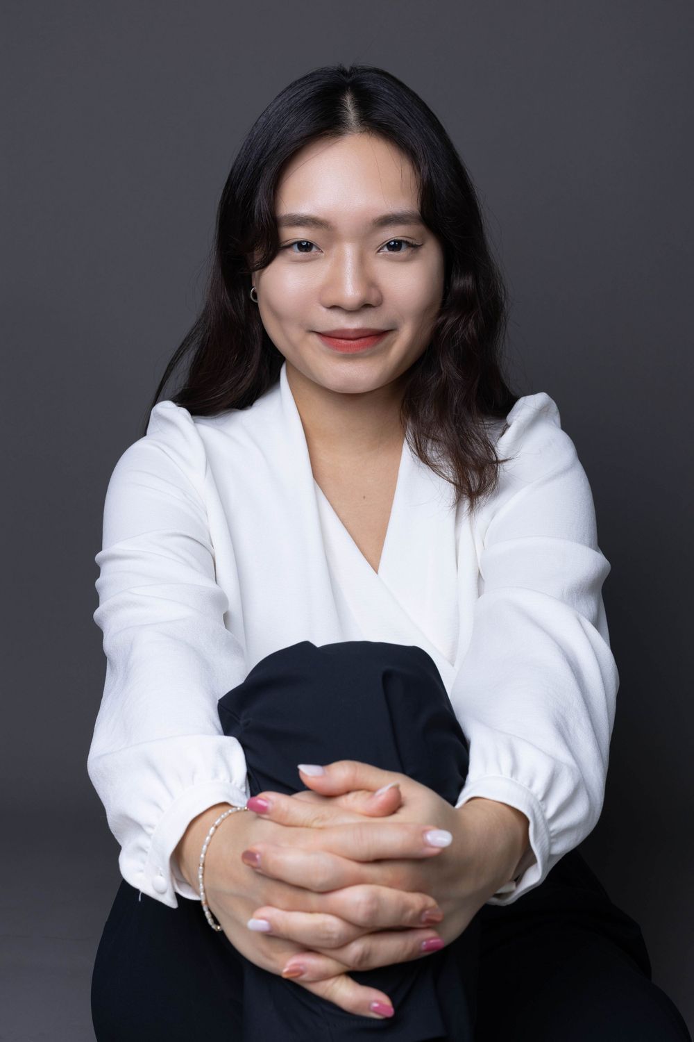 Asian woman in white blouse sitting with hands clasped smiling in professional studio portrait.
