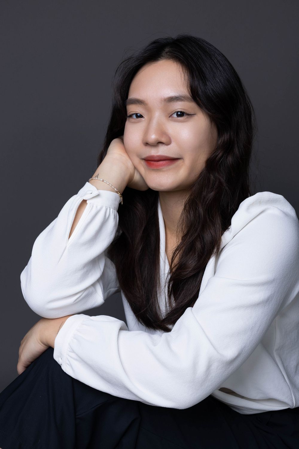 Relaxed Asian woman in white blouse sitting with hand on cheek smiling in studio portrait.