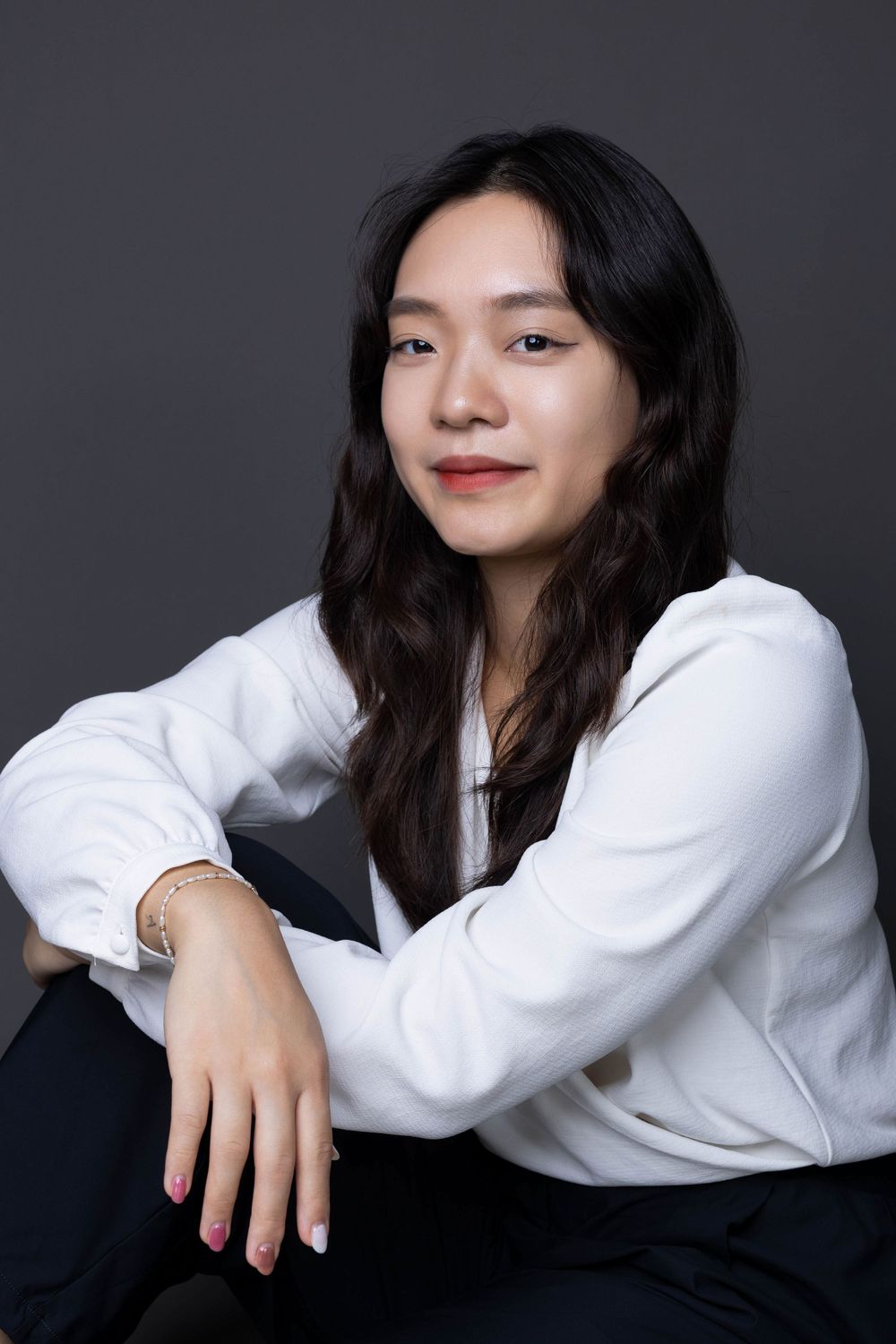 Asian woman in white blouse sitting casually with arms resting on knees in studio portrait.