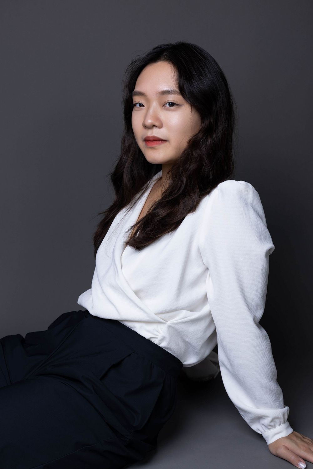 Young Asian woman in white blouse and black pants sitting gracefully against a dark gray studio background.