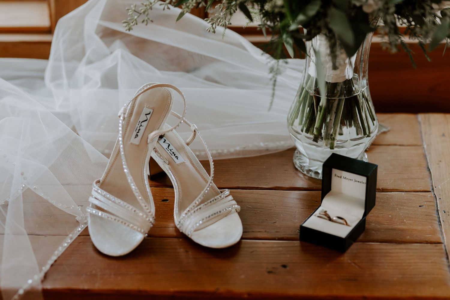 Wedding accessories including white heels, veil, and ring box arranged on wooden table.