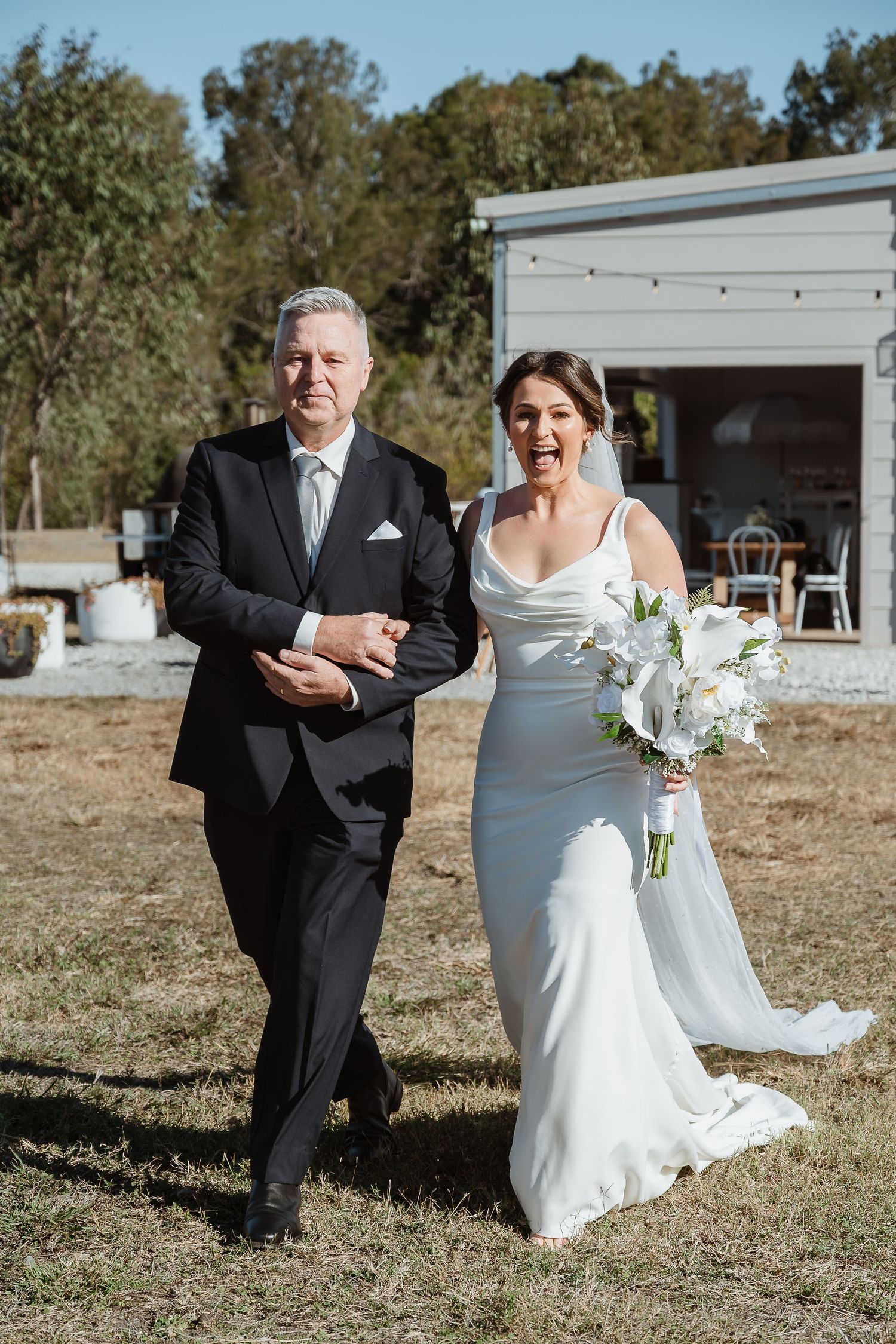 A bride in a flowing white dress walks through a lush green garden path at Yeppoon wedding venue on a sunny afternoon.