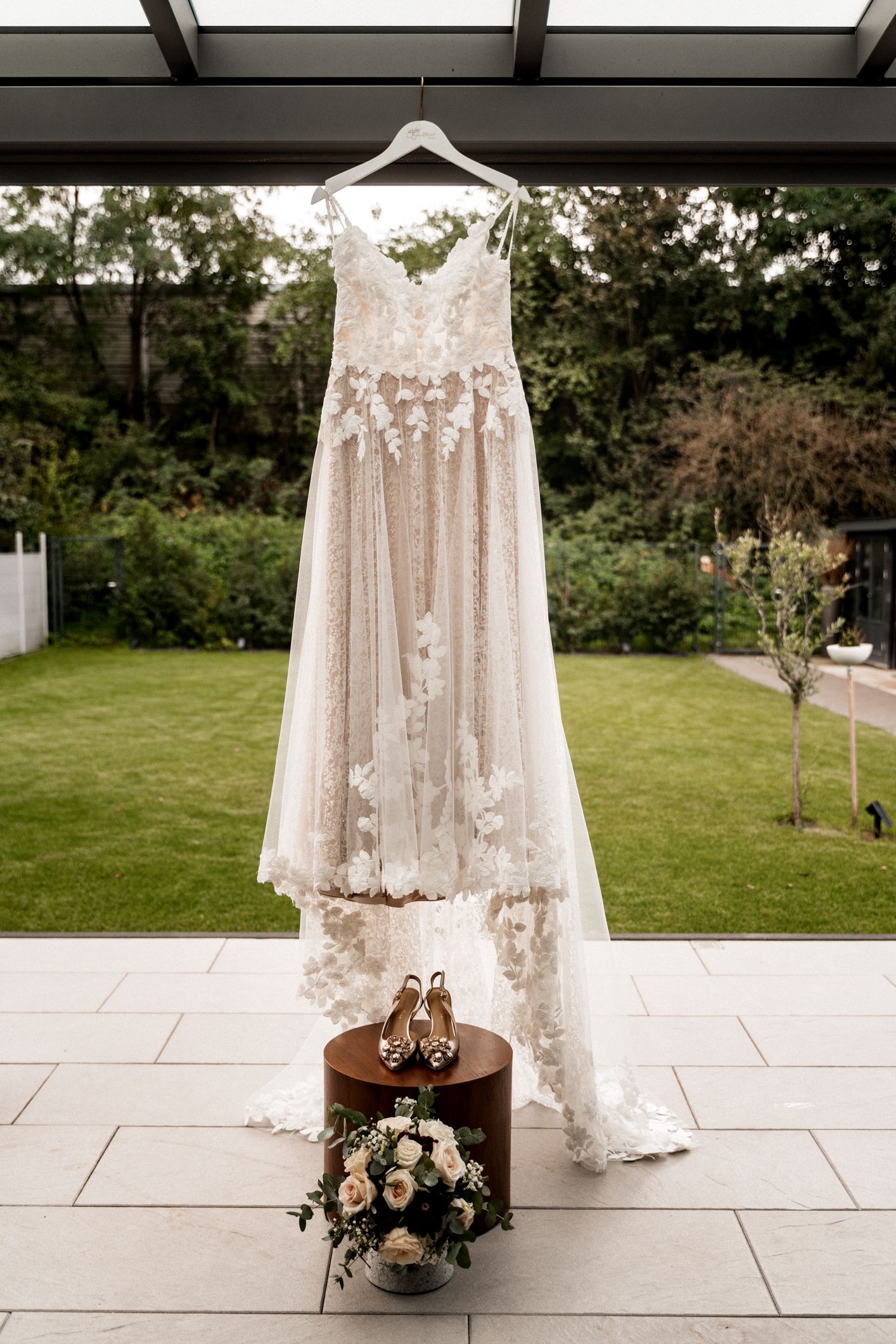 Elegant white lace wedding dress hanging with bridal shoes and floral bouquet on outdoor patio.