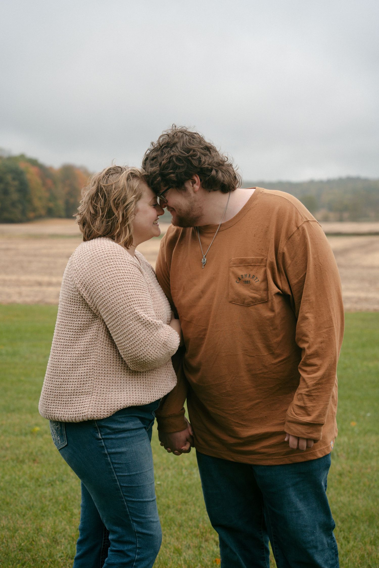 Couple holds hands and touches foreheads in an open field on a cloudy autumn day.