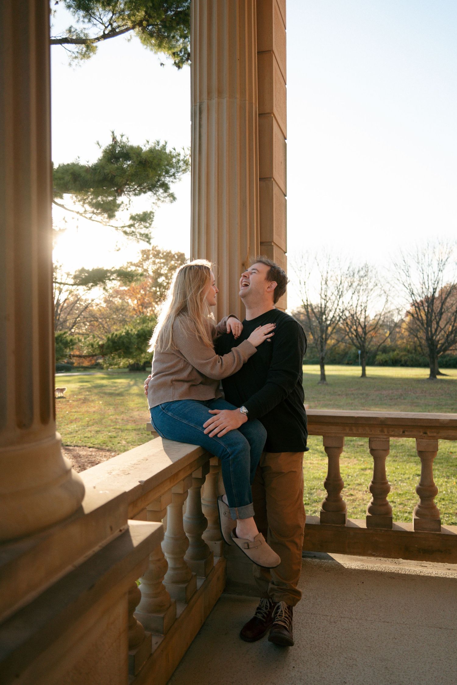Laughing couple embraces on stone balustrade of classical building in warm golden sunset light.