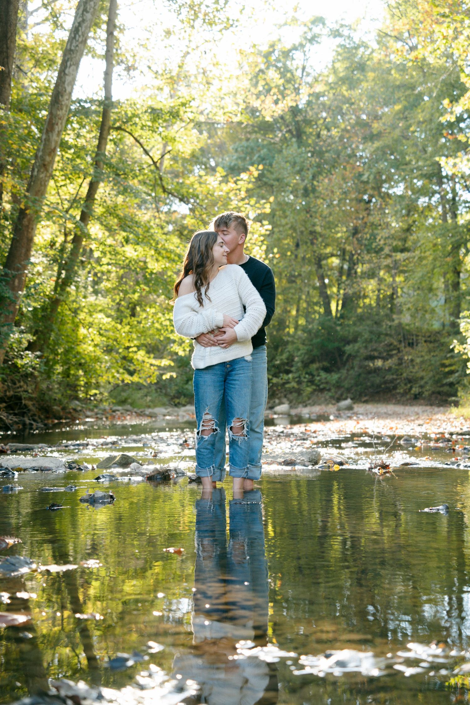 Couple kisses while standing in a shallow reflective creek surrounded by lush green forest.
