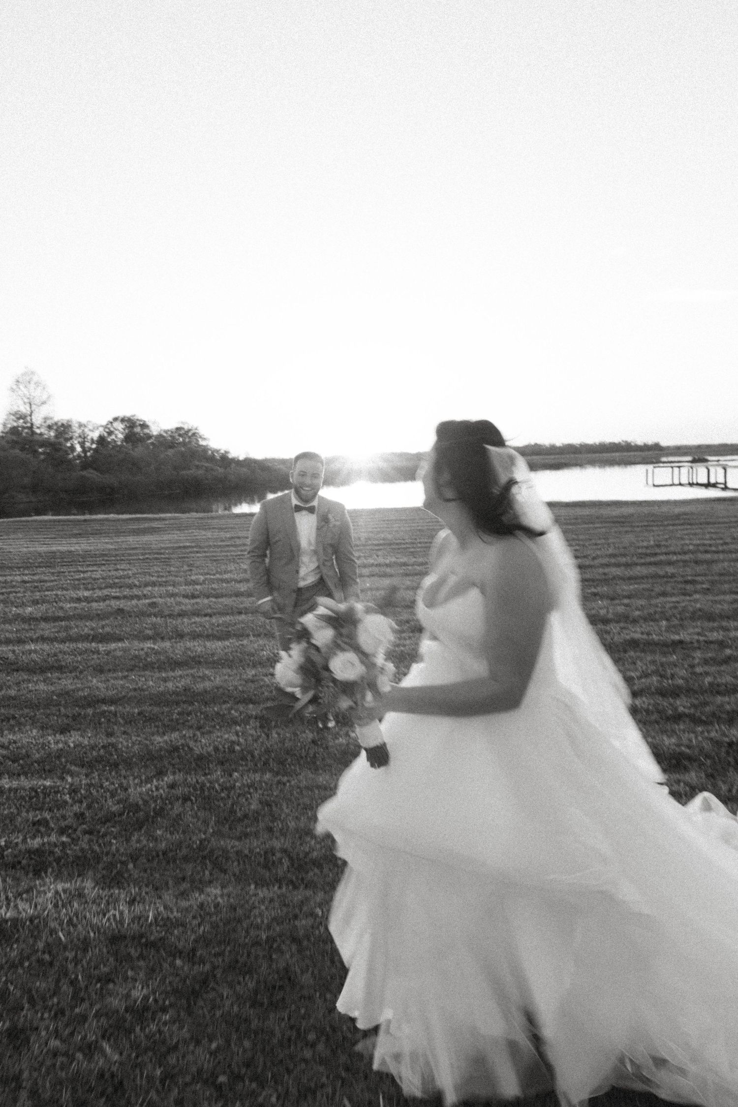 Black and white photo of bride in flowing dress standing in field at sunset.