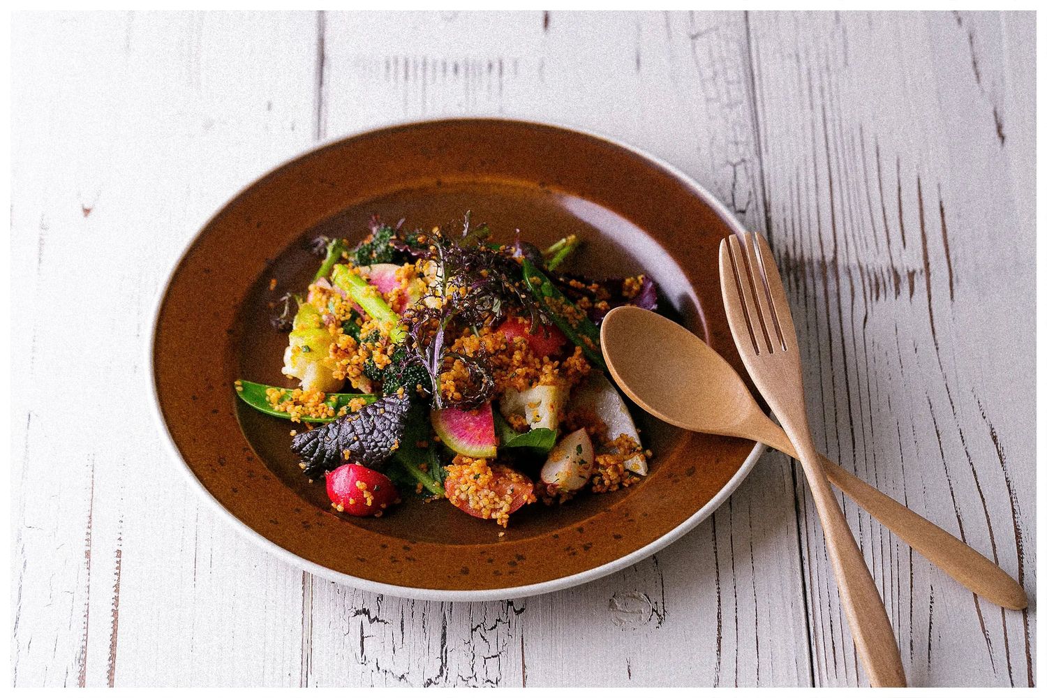 Colorful gourmet dishes plated on dark brown plates against a rustic white wooden background.