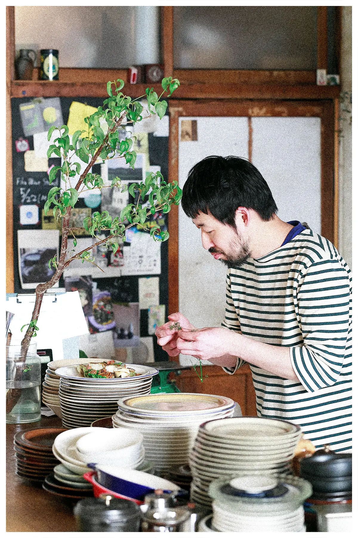 Person in striped shirt arranges stacks of white plates and dishes on wooden counter in rustic kitchen setting.