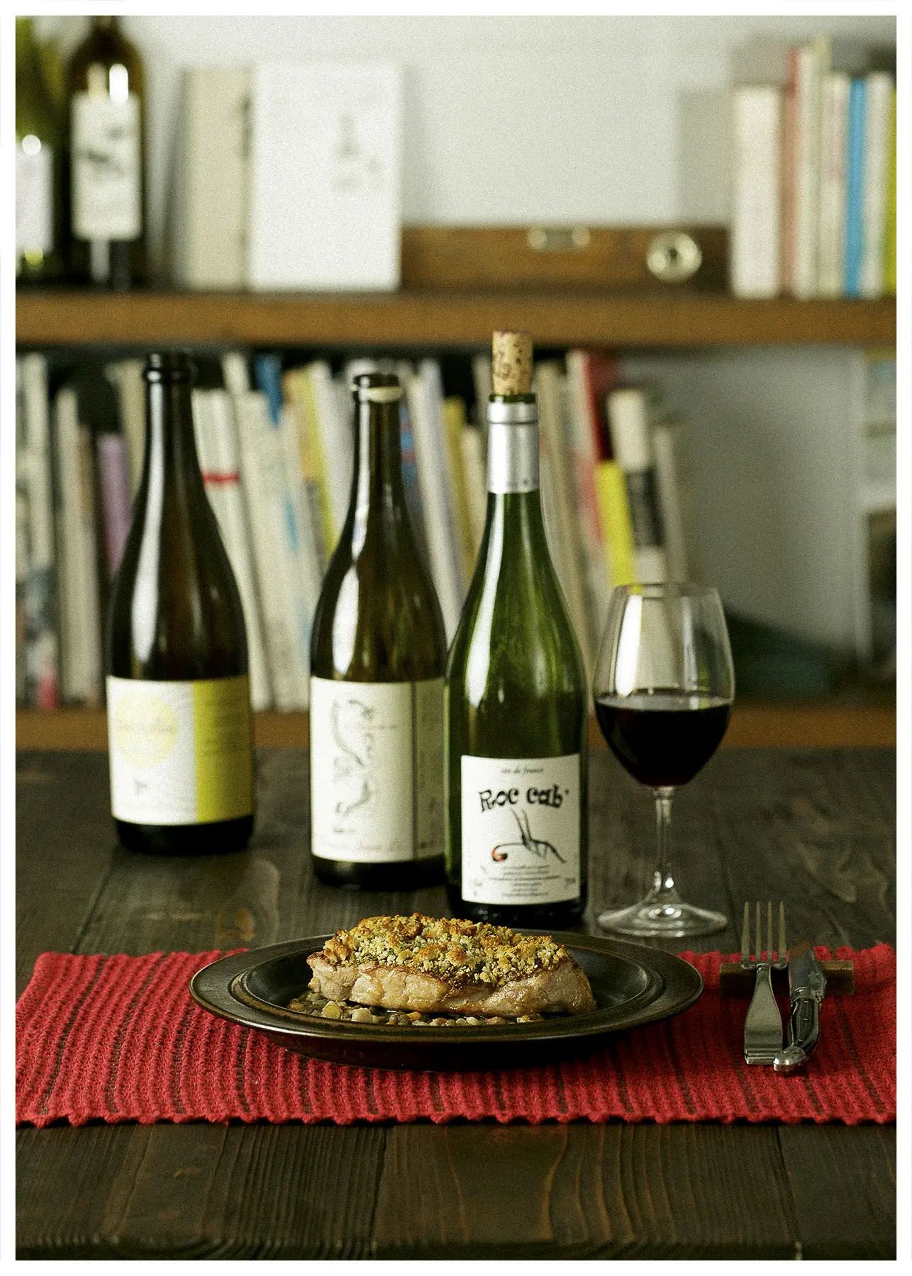 Wine bottles and glasses arranged on a dark wooden table with a red placemat and food plate create an elegant dining setup.