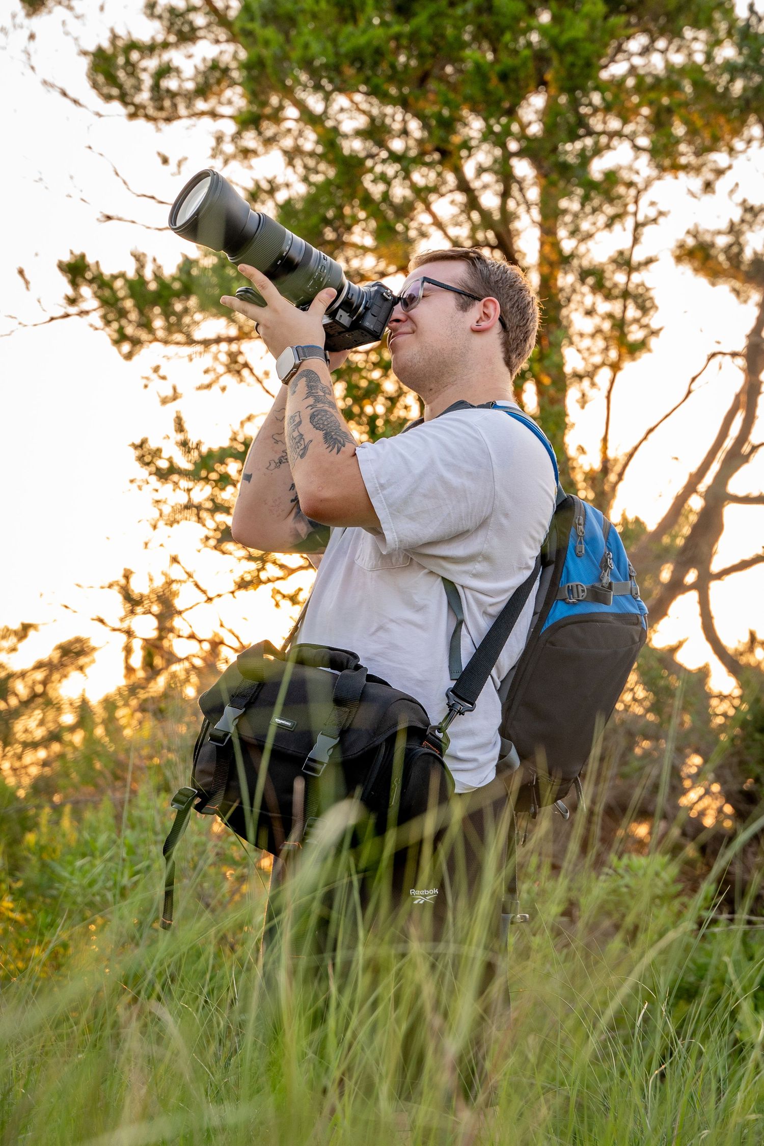 A photographer with a large telephoto lens takes photos in a grassy field at golden hour.