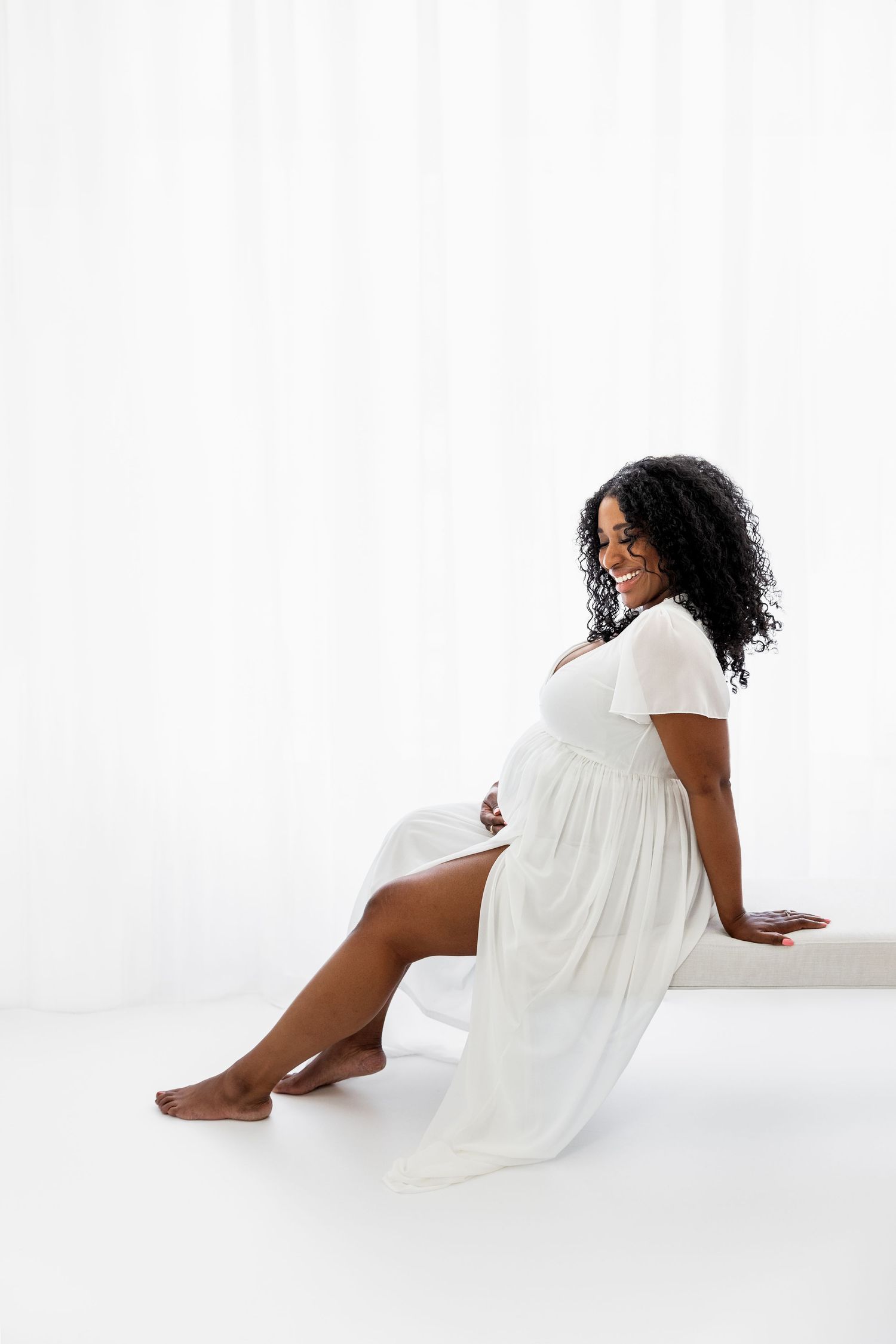 Gorgeous, confident black woman in white maternity dress, holding belly and smiling