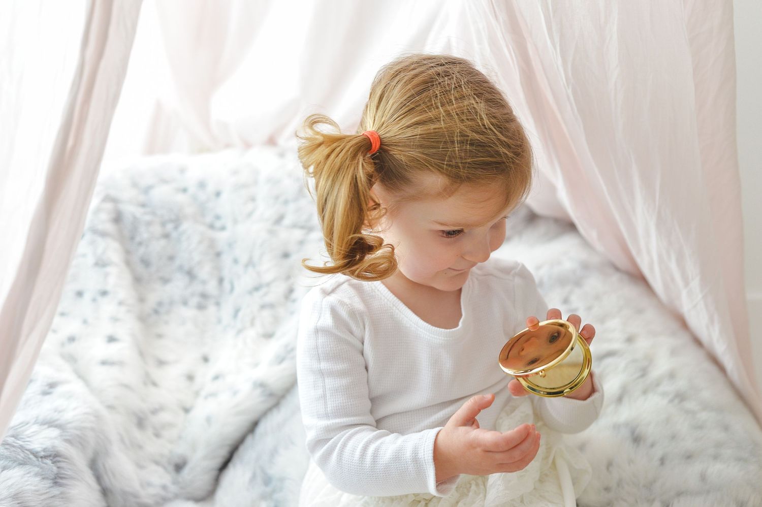 Young child in white shirt sits on bed holding compact mirror near sheer curtains in soft natural lighting.