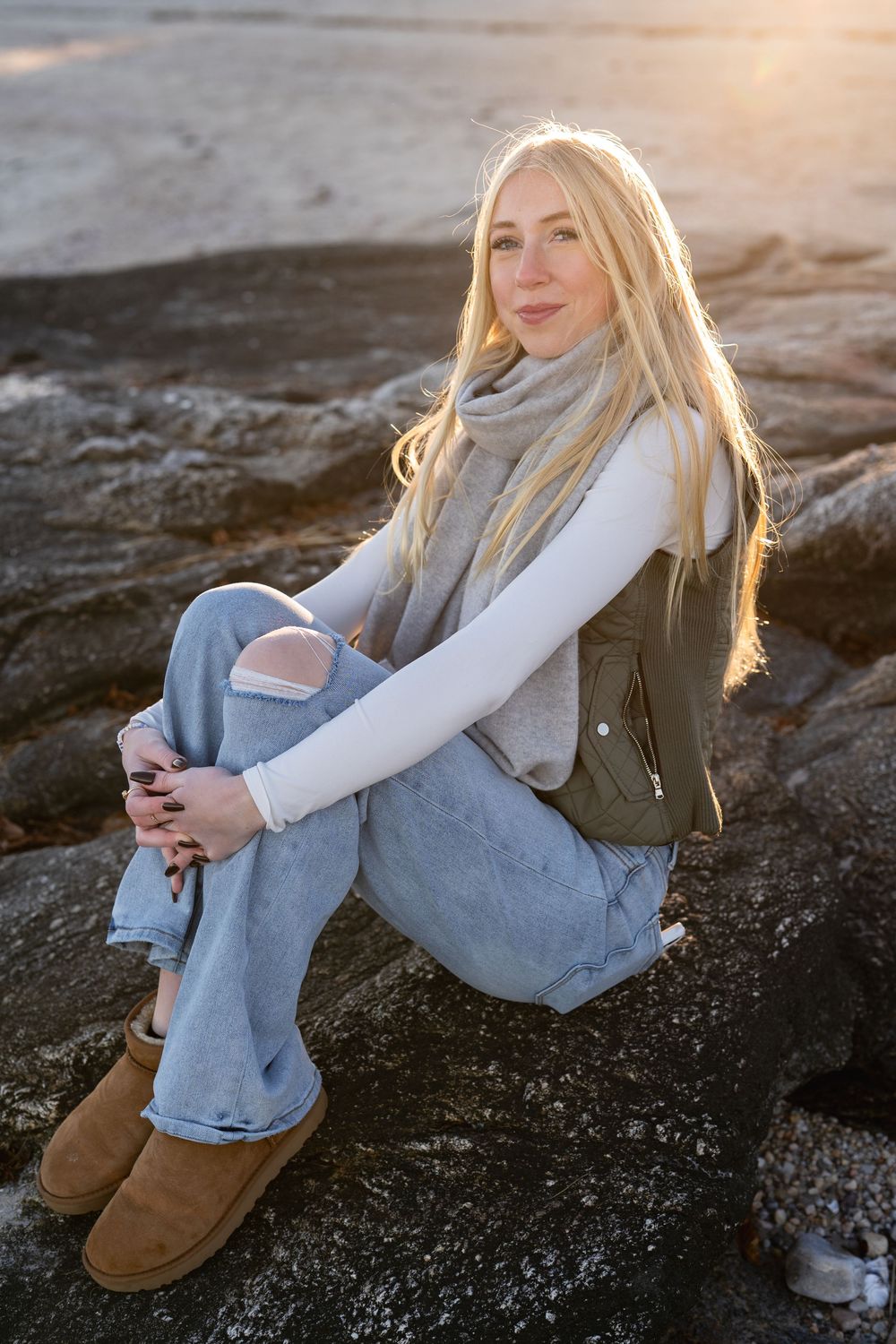 A person in denim overalls and white sweater poses on rocky terrain at sunset with golden backlighting.