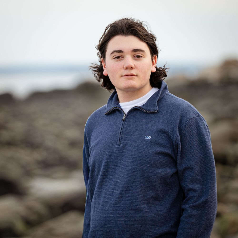 A high school senior boy in a navy Vineyard Vines pullover poses on rocky coastal shoreline with ocean in the background.