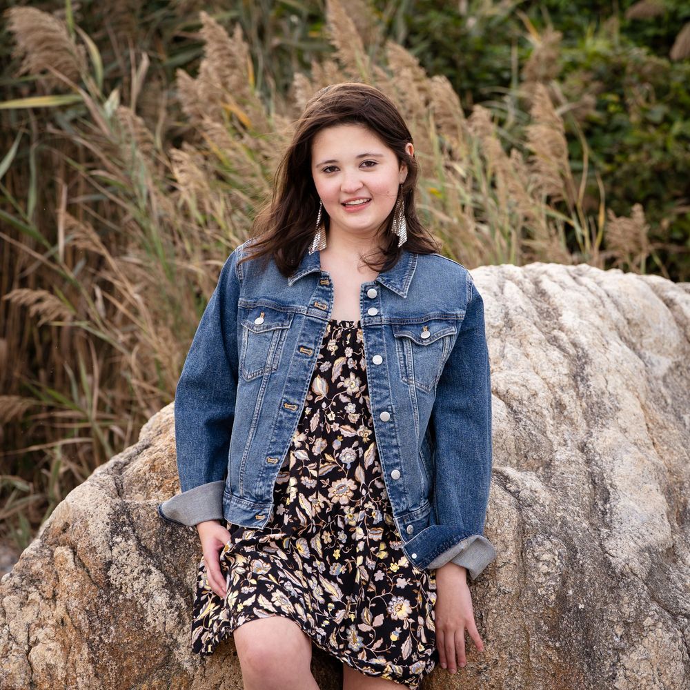 High school senior girl in denim jacket and floral dress poses on rocks with tall golden grasses in background.