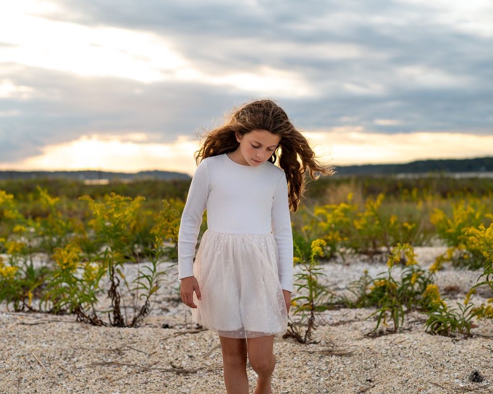 A white dress flows in the wind against a golden sunset backdrop over a sandy beach with scattered vegetation.
