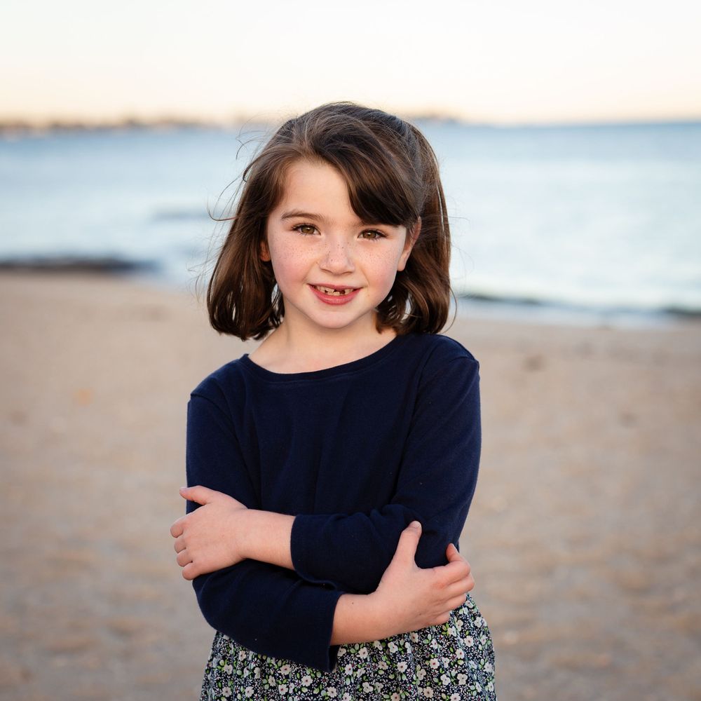 Young child in navy sweater and floral skirt poses with crossed arms at beach during sunset.
